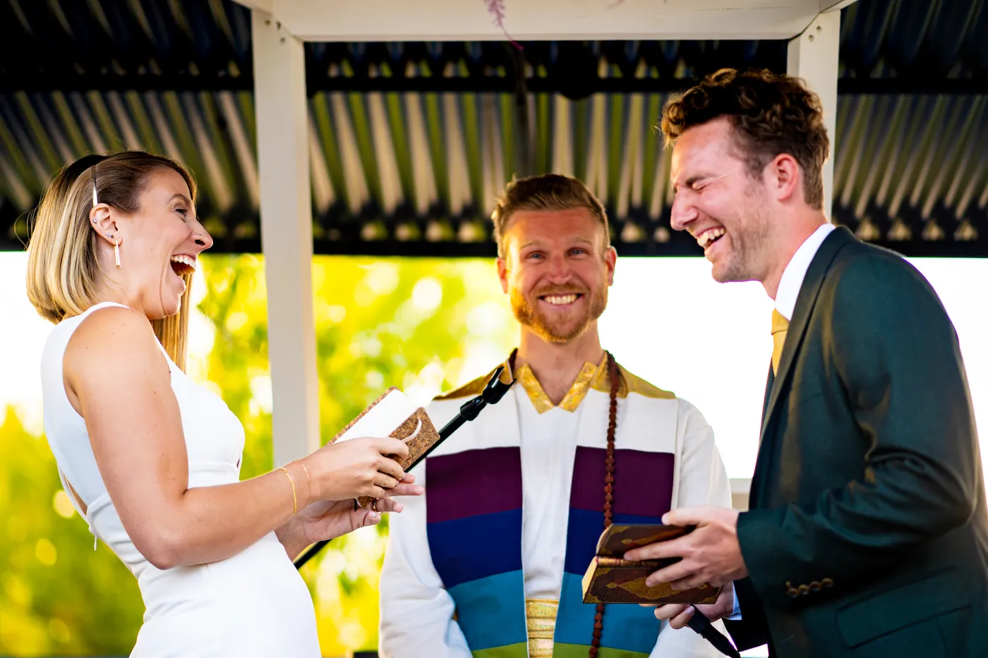 A couple laughing as they read their vows, with Telfer smiling between them in a rainbow stole.