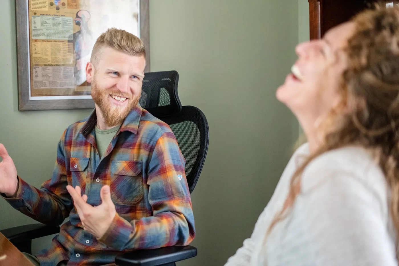 Telfer smiling and gesturing while a client laughs, a framed Ayurveda chart on the wall behind them.