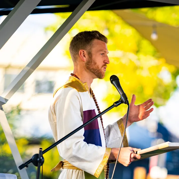 Telfer in a white and gold robe speaking into a microphone at an outdoor ceremony.