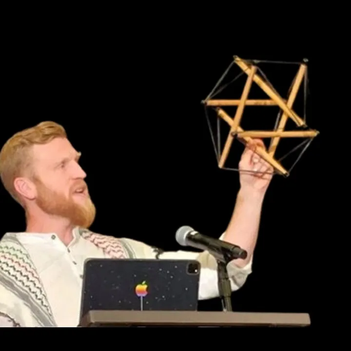 Telfer on a dark stage holding up a wooden tensegrity model beside a microphone.