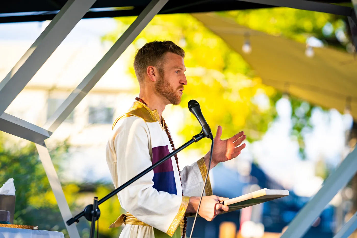 Telfer in white and gold robes speaking into a microphone under a canopy, autumn trees behind him.