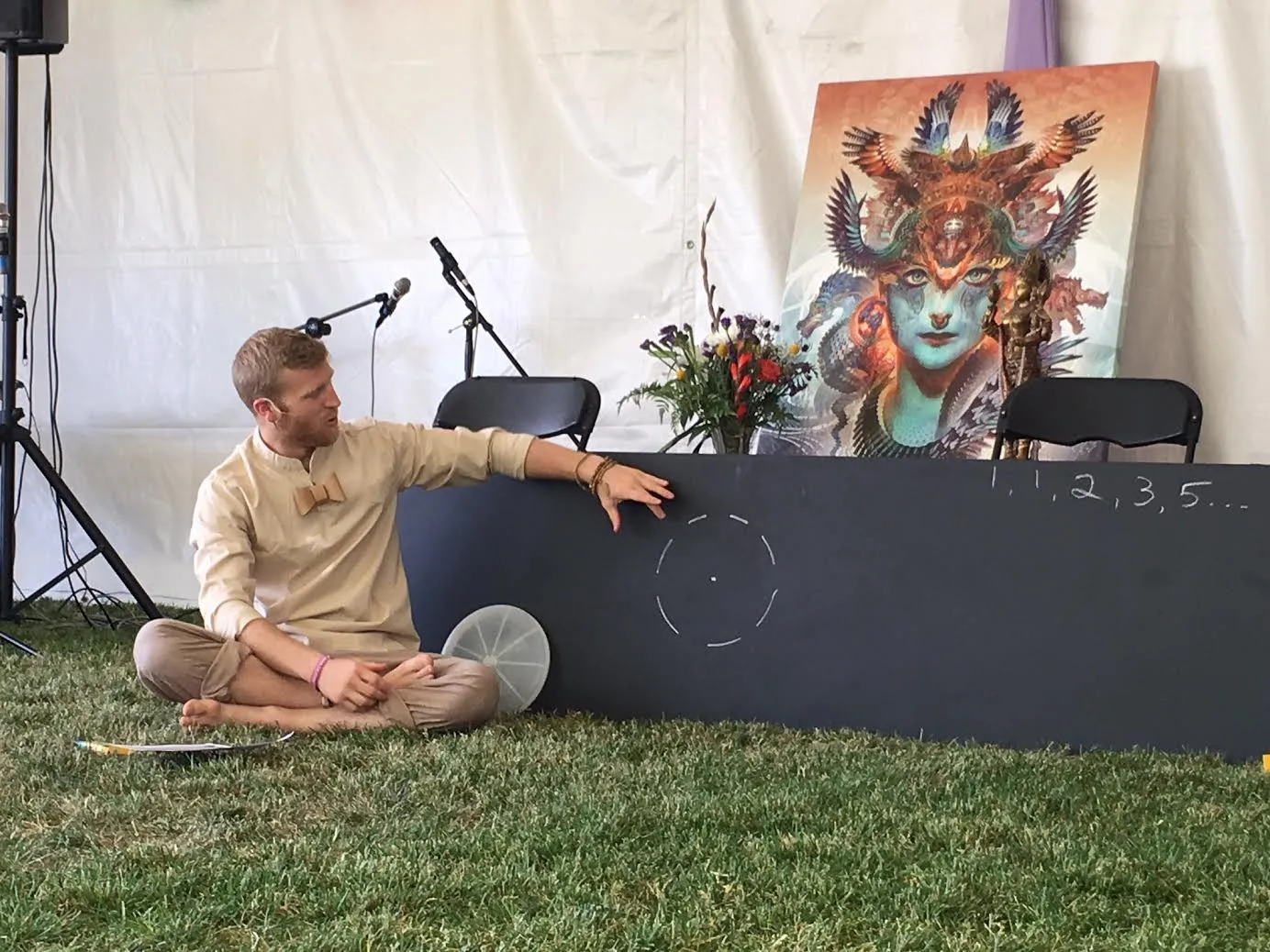 Telfer sitting cross-legged on the grass in a festival tent, pointing to a chalk circle and the Fibonacci sequence on a blackboard.