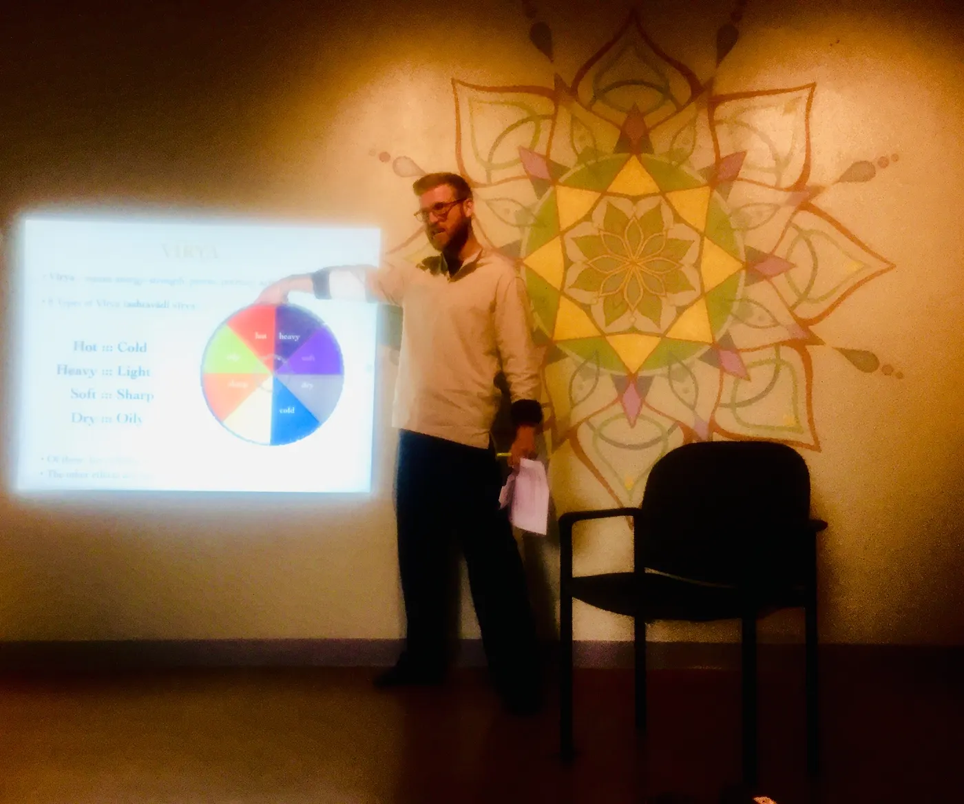 Telfer teaching in front of a projected slide on the eight qualities of virya, beside a painted mandala.